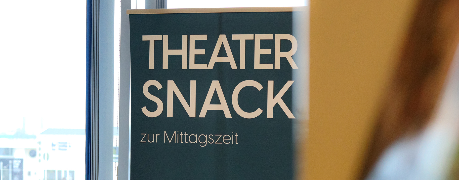 Petrolfarbenes Roll-up mit der Aufschrift "Theatersnack", im Vordergrund verschwommen ein Bücherregal aus seitlicher Perspektive