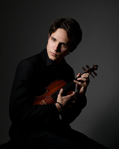 Portraitfoto von Javier Comesaña. Er sitzt seitlich und leicht nach vorn gebeugt. Er blickt zur Seite. In seinen Armen hält er eine Violine. Die Finger seiner linken Hand umfassen den Hals. Die Finger beider Hände zupfen über die Saiten. Er trägt ein schwarzes Hemd und eine schwarze Hose. Der Hintergrund ist dunkelgrau und indirekt ausgeleuchtet.