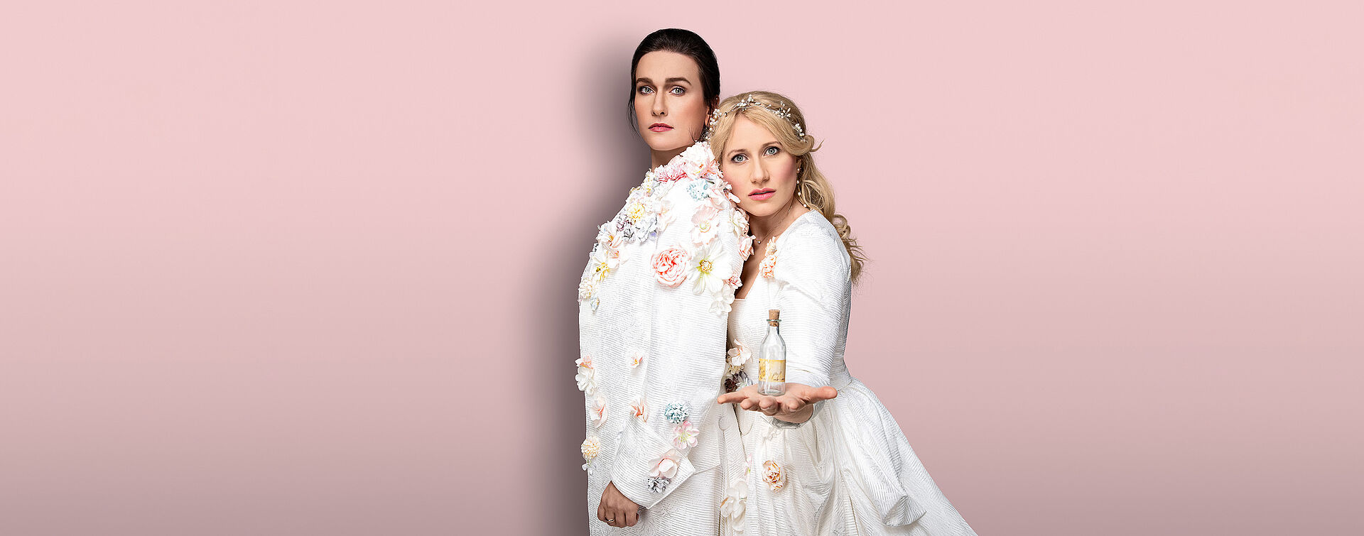 Zwei Frauen (Boshana Milkov und Victoria Kunze) stehen aneinander gelehnt. Boshana trägt eini männliches Gewand mit Blumen verziert. Ihre Haare hat sie nach hinten zusammengebunden. Victoria trägt ein weißen üppiges Kleid mit Blumen. Ihre blonden langen Haare fallen ihr in Locken über die Schulter. In ihrer Hand hält sie ein kleines Glasfläschchen. Der Hintergrund ist pastellrosa.