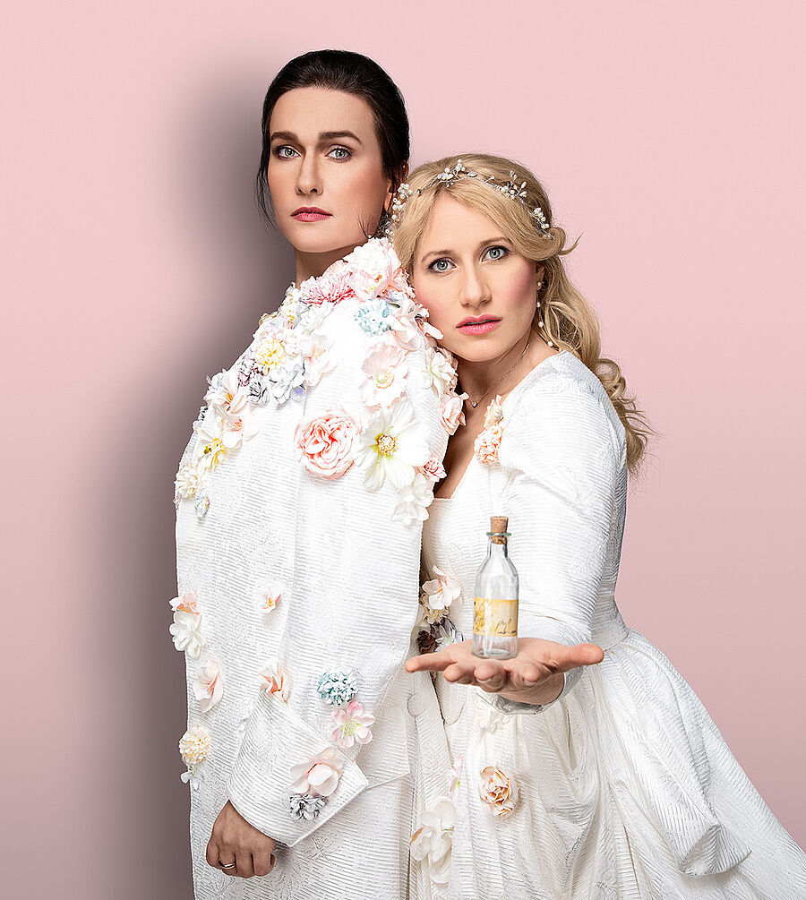 Zwei Frauen (Boshana Milkov und Victoria Kunze) stehen aneinander gelehnt. Boshana trägt eini männliches Gewand mit Blumen verziert. Ihre Haare hat sie nach hinten zusammengebunden. Victoria trägt ein weißen üppiges Kleid mit Blumen. Ihre blonden langen Haare fallen ihr in Locken über die Schulter. In ihrer Hand hält sie ein kleines Glasfläschchen. Der Hintergrund ist pastellrosa.