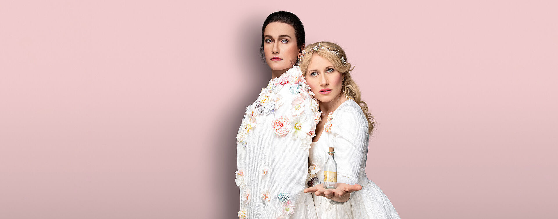 Zwei Frauen (Boshana Milkov und Victoria Kunze) stehen aneinander gelehnt. Boshana trägt eini männliches Gewand mit Blumen verziert. Ihre Haare hat sie nach hinten zusammengebunden. Victoria trägt ein weißen üppiges Kleid mit Blumen. Ihre blonden langen Haare fallen ihr in Locken über die Schulter. In ihrer Hand hält sie ein kleines Glasfläschchen. Der Hintergrund ist pastellrosa.