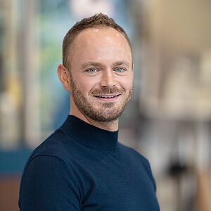 Portraitfoto von Markus Tatzig. Er trägt einen dunkelblauen Rollkragenpullover. Sein Haar ist braun und hochgegelt. Er trägt einen Dreitagebart. 