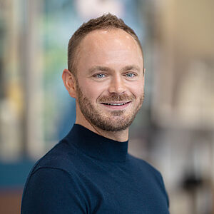 Portraitfoto von Markus Tatzig. Er trägt einen dunkelblauen Rollkragenpullover. Sein Haar ist braun und hochgegelt. Er trägt einen Dreitagebart. 