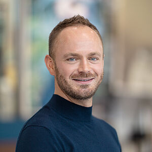 Portraitfoto von Markus Tatzig. Er trägt einen dunkelblauen Rollkragenpullover. Sein Haar ist braun und hochgegelt. Er trägt einen Dreitagebart. 