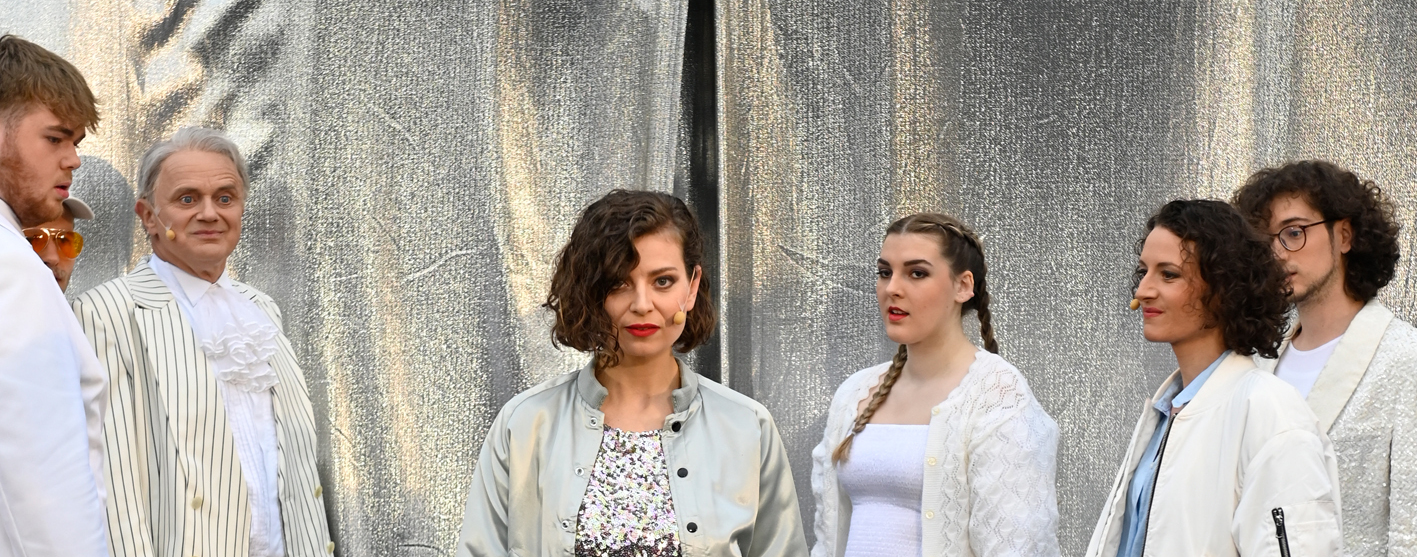 Roxane schreitet mit dem Blick aufs Publikum gerichtet aufs selbige zu. Dabei trägt sie ein glitzerndes Kleid, welches den silbernen Stoff im Hintergrund wiederspeigelt. Das Ensemble bildet eine Art Korridor um Roxane und schauen ihr hinterher.
