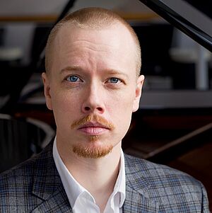 Portraitfoto von Bryan Boyce. Er trägt sein blondes Haar kurzgeschoren, dazu einen Schurr- und Kinnbart. Er ist in ein kariertes blau-graues Jackett gekleidet. Darunter trägt er ein weißes Hemd. Er sitzt vor einem Flügel.