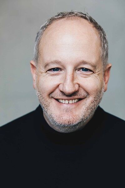 Portraitfoto von Achim Lenz. Er grinst mit offenem Mund. Er hat graues kurzes Haar und einen Dreitagebart. Er trägt einen schwarzes Pullover. Der Hintergrund ist hellgrau.