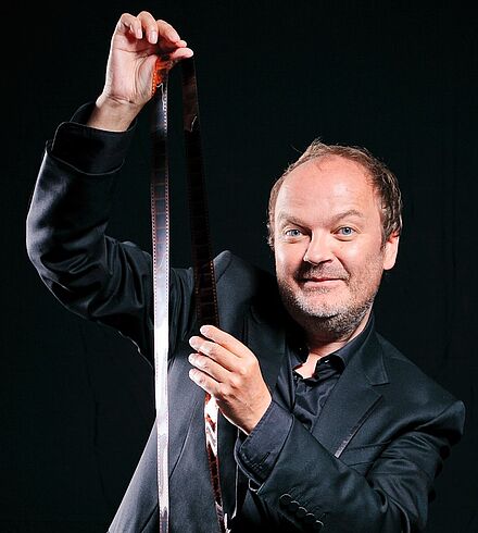 Jens Wawrczeck sitzt in schwarzem Anzug vor schwarzem Hintergrund und hält ein Filmband empor.