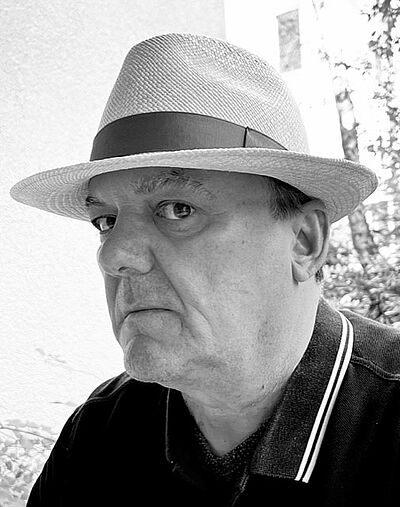 Schwarzweißes Portraitfoto von Stefan Mayer. Er trägt einen hellen Hut mit schmaler Krempe und schaut seitlich in die Kamera. Dazu trägt er ein dunkles Poloshirt. im hintergrund sind Bäume zu erahnen.
