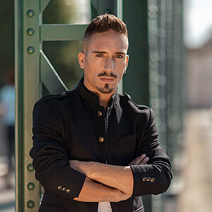 Portraitfoto von Stefano Neri. Er lehnt mit verschränkten Armen an grünen Stahlstreben. Er trägt eine schwarze Jacke, darunter ein weißes T-Shirt. Sein Haar ist hochgegelt.