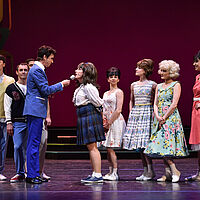 Tracy Turnblad spricht in ein Mikrofon, das Corny Collins ihr hinhält. Sie blicken einander an. Einige Corny Collins Kids schauen zu. Tracy Turnblad trägt eine Schuluniform, die anderen Kleider mit Petticoats und bunte Kostüme.