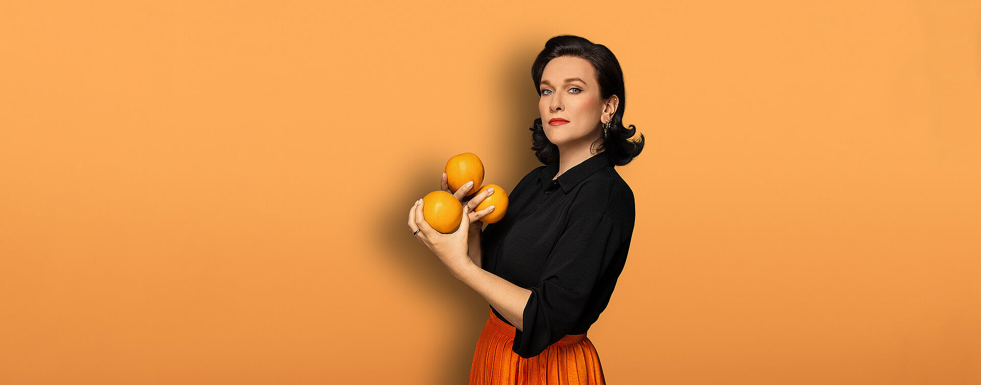 Eine Frau (Boshana Milkov) hat schulterlange schwarze Haare. Sie trägt eine schwarze Bluse und einen orangefarbenden Faltenrock. In ihren Händen hält sie drei Orangen. Der Hintergrund des Bildes ist orange.