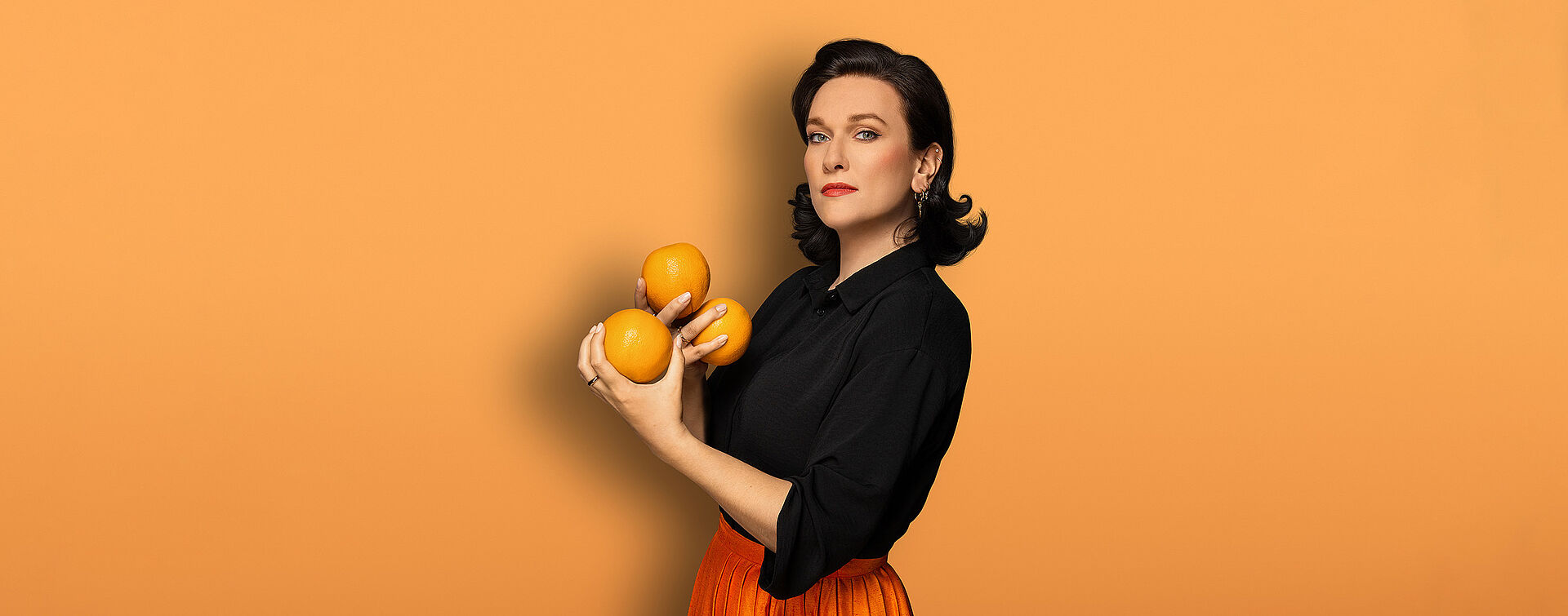 Eine Frau (Boshana Milkov) hat schulterlange schwarze Haare. Sie trägt eine schwarze Bluse und einen orangefarbenden Faltenrock. In ihren Händen hält sie drei Orangen. Der Hintergrund des Bildes ist orange.