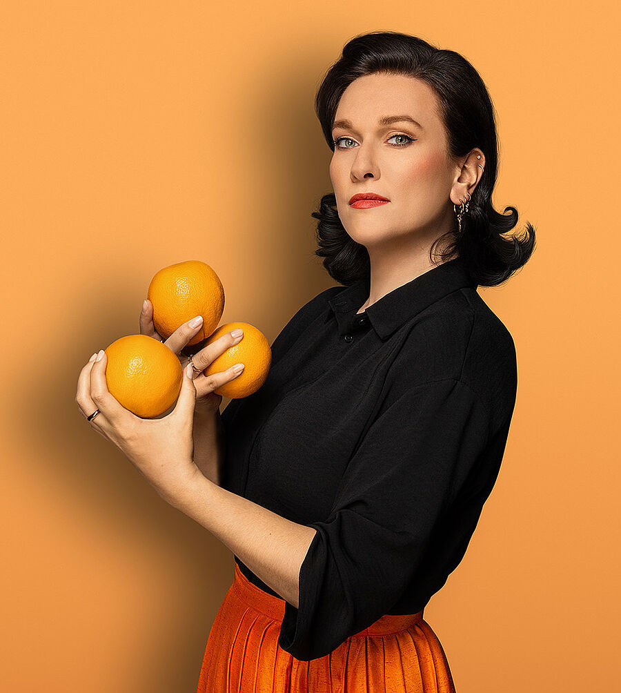 Eine Frau (Boshana Milkov) hat schulterlange schwarze Haare. Sie trägt eine schwarze Bluse und einen orangefarbenden Faltenrock. In ihren Händen hält sie drei Orangen. Der Hintergrund des Bildes ist orange.