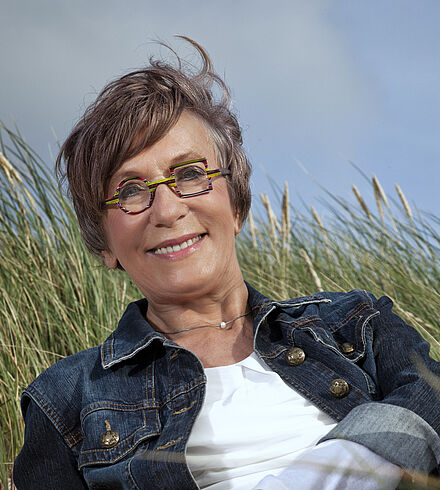Portraitfoto von Gisa Pauly. Sie grinst in die Kamera. Ihr kurzes Haar wehnt im Wind. Sie trägt eine bunte Brille, dessen eines Glas rund und das andere quadratisch ist. Im Hintergrund weht Schilfrohr. Der Himmel ist blau-grau.