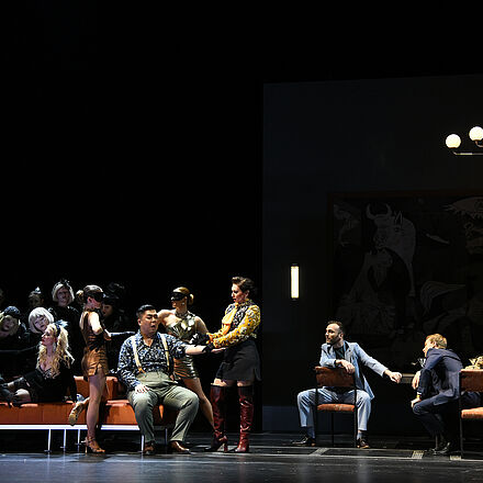 Zu erkennen ist eine große Gruppe von Menschen. Im Mittelpunkt sitzt ein Mann mit ausgestrecktem Arm, an dem eine Frau eine Spritze ansetzt. Rechts im Bild sitzen zwei Männer an einem Tisch und beobachten die Situation.