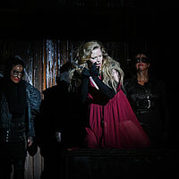 Lady Macbeth hält ein Messer in der Hand und beugt sich singend nach vorn. Hinter ihr sind die Hexen aufgereiht.