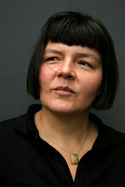 Portraitfoto von Susana Mendoza. Sie steht vor einer dunkelgrauen Wand. Sie ist schwarz gekleidet. Sie trägt eine Bluse und eine Kette. Sie trägt kinnlanges Haar und einen Pony.