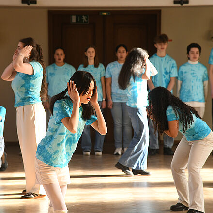 Im Vordergrund sind mehrere Kinder zu sehen, die sich Augen, Ohren oder Mund zuhalten. Im Hintergrund steht eine Reihe von Kindern. Alle tragen ein blaues Oberteil.