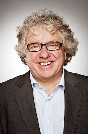 Portraitfoto von Christoph Nix. Er steht grinsend vor einem grauen Hintergrund. Er hat graues Haar, trägt eine schwarze Brille und ein dunkles Jackett zu hellblauem Hemd.