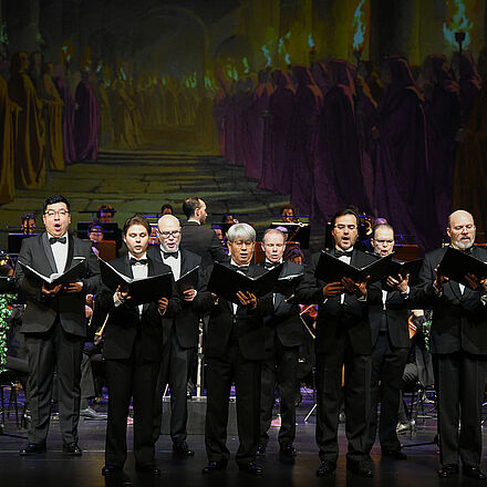 Der Herrenopernchor steht mit Noten in der Hand vor dem Orchester.  