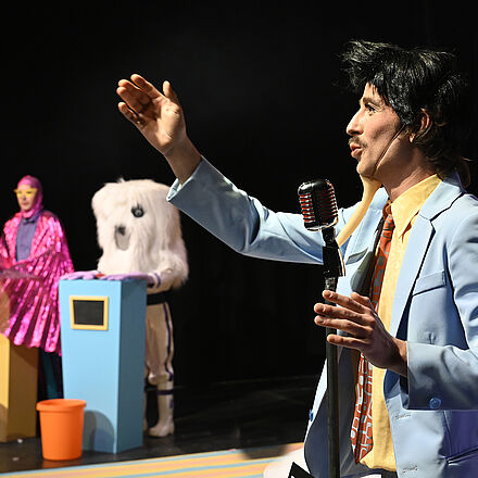 Im Vordergrund steht am Mikro und zeigt ins Publikum. Im Hintergrund stehen zwei Personen hinter jeweils einem Pult. 