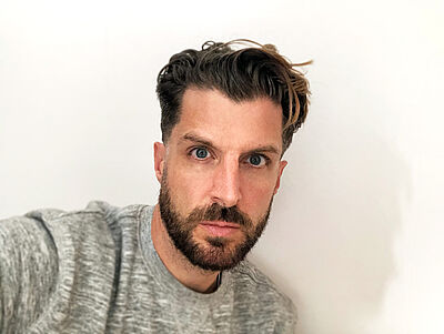 Selfie von Alex Lowde. Er hat braunes Haar, dass an den Seiten kurz ist. Die obere Partie ist locker zurückgegelt. Er hat einen Vollbart. Er trägt einen grau-melierten Pullover und steht vor einer weißen Wand. Der Schatten seines Gesichtes fällt darauf.