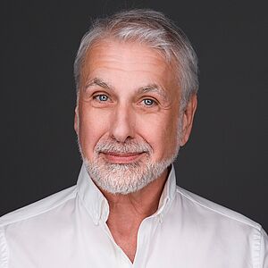 Portraitfoto von Hans Neblung. Er lächelt verschmitzt. Sein weißes Haar trägt er kurz, dazu einen kurzen Vollbart. Er ist in ein weißes Hemd gekleidet. Der Hintergrund ist dunkelgrau.