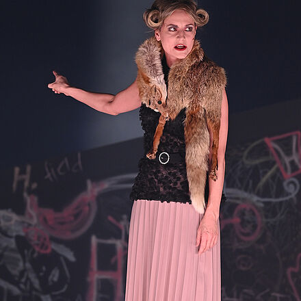 Im Vordergrund steht eine Frau, die einen Fuchsschal umgebunden hat. Sie zeigt in den Hintergrund. Dort befindet sich eine mit Graffiti besprühte Wand.