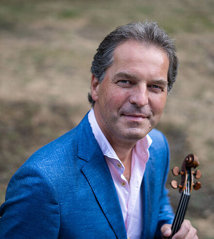 Portraitfoto von Frank-Michael Erben. Er trägt ein helles Hemd und en blaues Sakko. In seiner linken Hand hält er eine Violine. Er hat kurze dunkle Haare, die nach hinten gekämmt sind. 