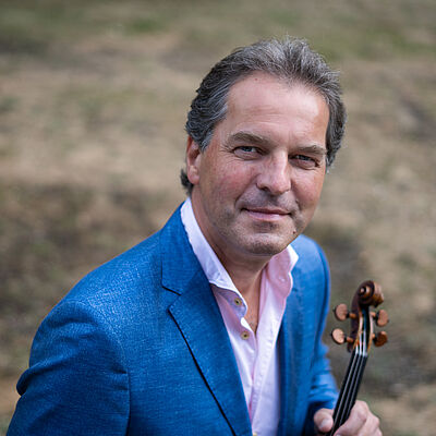 Portraitfoto von Frank-Michael Erben. Er trägt ein helles Hemd und en blaues Sakko. In seiner linken Hand hält er eine Violine. Er hat kurze dunkle Haare, die nach hinten gekämmt sind.