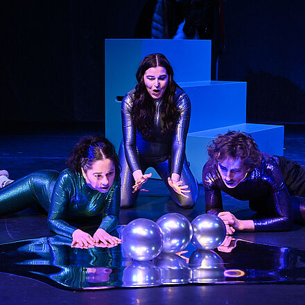 Die drei Personen schauen fragend auf drei Ballons, die sich auf dem Boden spiegeln.