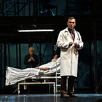 Dr. Richardson steht im Arztkittel vorn. Im Hintergrund sitzt Bess McNeill am Krankenbett von Jan Nyman.