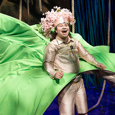 Eine Person mit Blumen auf dem Kopf und einem grünen Umhang lacht.