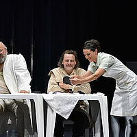 Dorfrichter Adam und Schreiber Licht sitzen an einem Tisch. Marthe Rull steht neben Schreiber Licht und deutet auf eine Scherbe in ihren Händen. Sie blicken gemeinsam auf diese und grinsen. Dorfrichter Adam blickt mit verkniffenem Blick herüber. Die Kleidung und die Einrichtung sind weiß. Dorfrichter trägt eine Beule auf der Stirn.