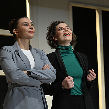 Eine Frau im grauen Anzug steht neben der Frau im schwarzen Anzug. Beide blicken siegessicher.