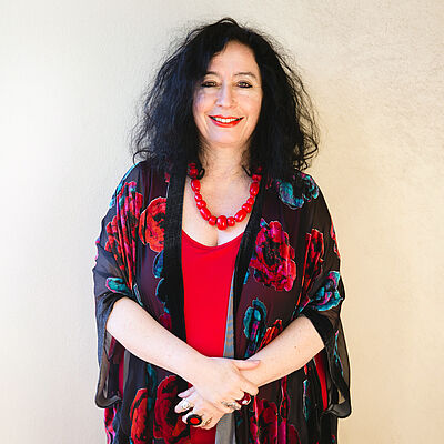 Portraitfoto von Elena Kats-Chernin. Ihr schwarzes Haar fällt ihr auf die Schultern. Sie ist in ein rotes Oberteil gekleidet, darüber trägt sie einen geblümten Poncho. Auf ihrem Dekoltee liegt eine rote Perlenkette auf. Ihr Hände sind vor dem Bauch übereinander gelegt. Sie grinst mit roten Lippen.