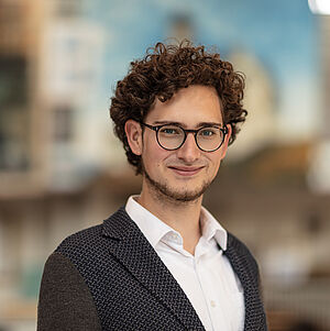 Portraitfoto von Victor Seraphin Feuchte. Er hat lockiges dunkles Haar, eine runde schwarze Brille und einen gesichtsrahmenden Bart. Er trägt ein weißes Hemd, darüber ein blau-graues Jackett.