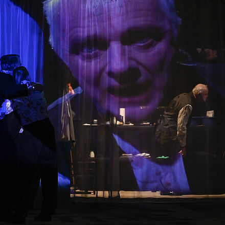 Im Vordergrund hängt ein Vorhang, auf den ein Gesicht projiziert ist. Im Hintergrund, hinter dem Vorhang, stehen drei Menschen.