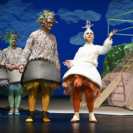 Eine Gans zeigt Richtung Himmel. Links neben ihr steht eine Wildgans. Im Hintergrund sieht man Wolken, ein Podest aus Papier und weitere Wildgänse