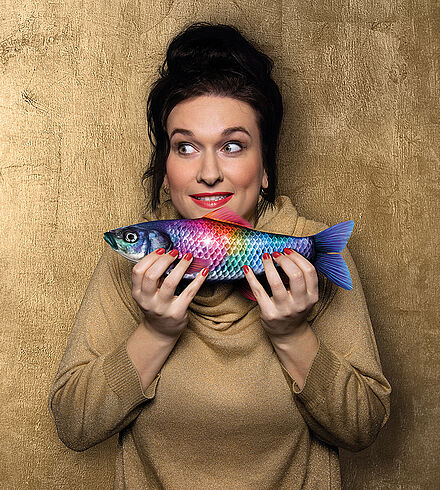 Frau in goldenem Kleid hält einen bunten Fisch