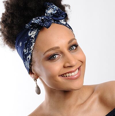 Portraitfoto von Vanessa Weiskopf. Sie hat ihren Kopf seitlich gedreht. Ihr lockiges dunkles Haar ist zu einem lockeren Dutt gebunden, welcher von einem blau-weißen Haarband mit Schleife eingefasst ist. Sie lächelt mit geöffnetem Mund. In ihrem Ohr trägt sie tränenförmige Ohrringe. Sie trägt ein schulterfreies schwarzes Oberteil. Der Hintergrund ist weiß.