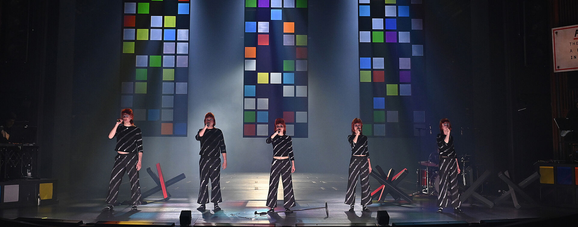 Fünf gleich gekleidete Personen stehen nebeneinander in einer Reihe und halten Mikrofone in der rechten Hand. Im Hintergrund schweben drei bunte Glaswände in der Luft. Auf dem Boden stehen mehrere Grenzblockaden.