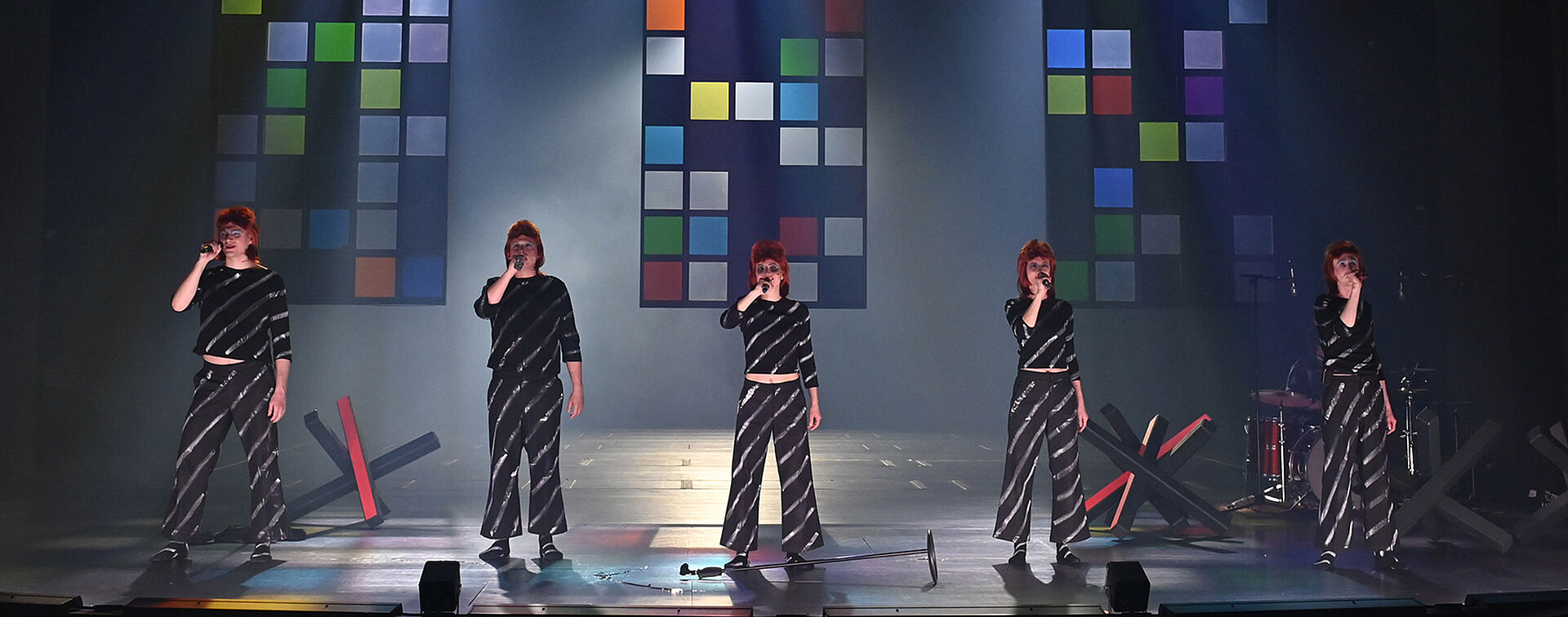 Fünf gleich gekleidete Personen stehen nebeneinander in einer Reihe und halten Mikrofone in der rechten Hand. Im Hintergrund schweben drei bunte Glaswände in der Luft. Auf dem Boden stehen mehrere Grenzblockaden.