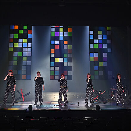 Fünf gleich gekleidete Personen stehen nebeneinander in einer Reihe und halten Mikrofone in der rechten Hand. Im Hintergrund schweben drei bunte Glaswände in der Luft. Auf dem Boden stehen mehrere Grenzblockaden.
