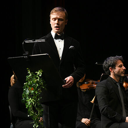 Im Vordergrund singt eine Person, während im Hintergrund das Orchester sitzt.  