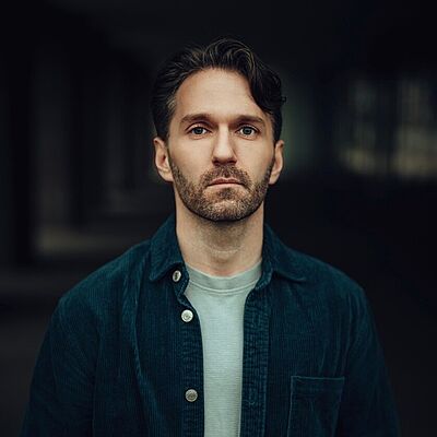 Portraitfoto von Stefan Preuth. Er hat kurze dunkle Haare, die ihm in die Stirn fallen und einen Bart. Er trägt ein hellblaues T-Shirt und darüber ein offenes dunkelblaues Hemd. Er schaut ernst.