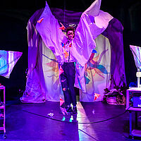 Zwei Personen in ein Bettlaken gewickelt, umsäumen einen Mann, der seine Arme in Bettlaken emporhält. Die Bühne ist violett ausgeleuchtet. Links und rechts steht je ein Overhead-Projektor mit einer gläsernen, mit Wasser gefüllten Auflaufform.