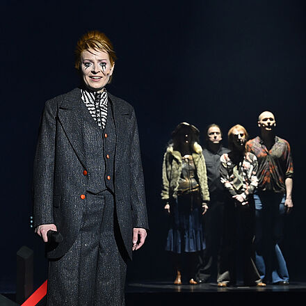 Im Vordergrund steht eine Frau im Anzug mit schwarz unterlaufenen Augen. Hinter ihr befindet sich eine Grenzblockade. Rechts im Hintergrund stehen vier Personen.