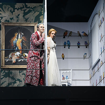 In der Mitte des Bildes stehen links ein Mann und rechts eine Frau nebeneinander. Zwischen den beiden Personen befindet sich eine Tür.